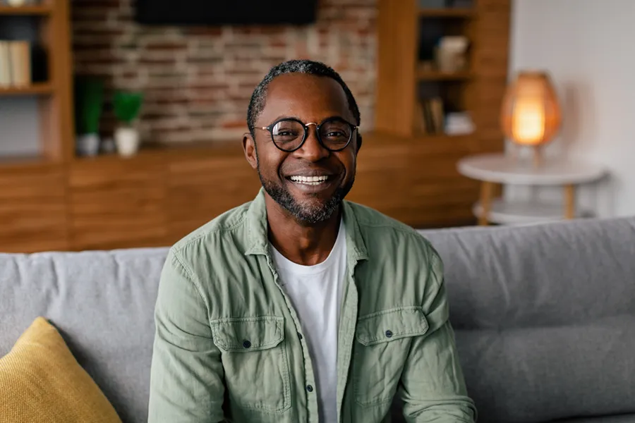 A middle aged black man in a green button up shirt sits on his couch smiling, happy with his treatment for andropause and low testosterone from Raving Health in Virginia Beach.