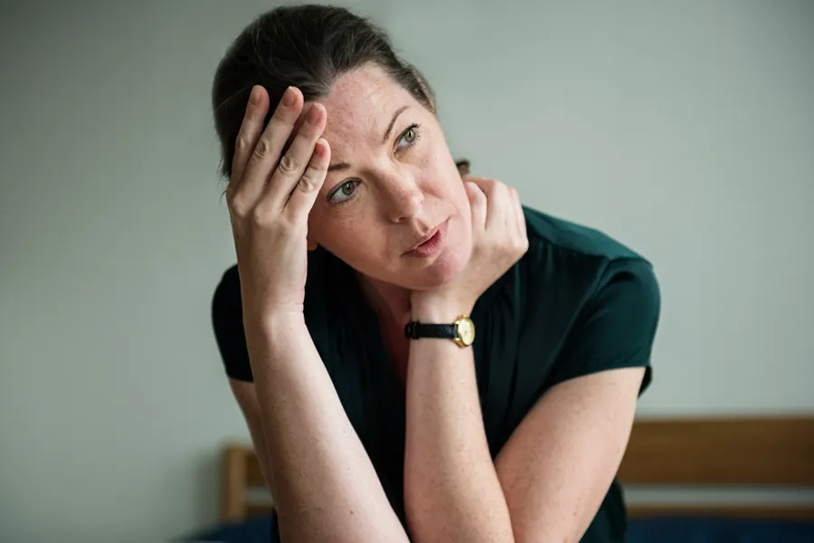 A woman in a dark blue dress sits on her bed holding her head in her hands with an exhausted expression. Get help for chronic fatigue syndrome from Raving Health in [AERA].