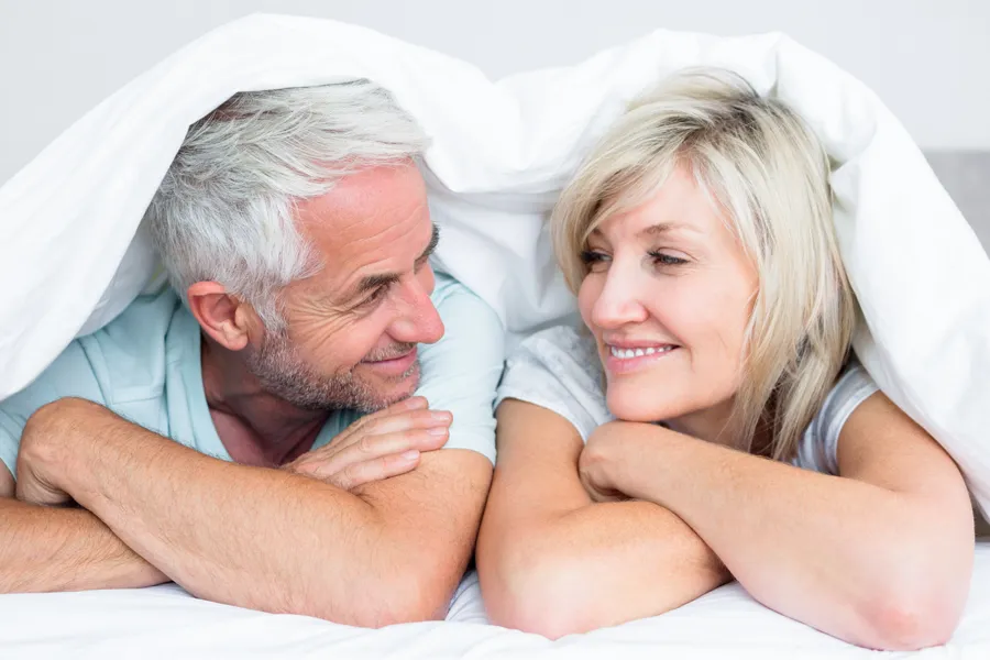 A mature couple lay under their bed's covers and smile at each other. Get treatment for low libido from Raving Health in Virginia Beach.
