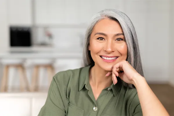 Menopause-Clinic An asian woman with long gray hair and a green shirt sitting and smiling on her couch, happy with her menopause care from [NAME} in Virginia Beach.