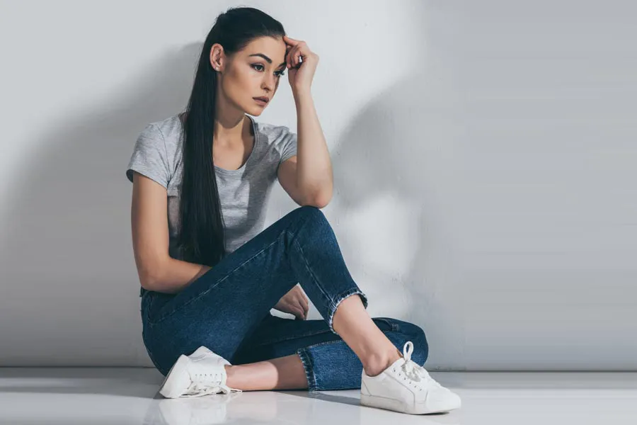 PCOS-Doctor A young brunette woman in a gray t-shirt and jeans sits against a gray wall with her head resting on her hand and a distressed expression. Get help for Polycystic Ovarian Syndrome (PCOS) from Raving Health in Virginia Beach.