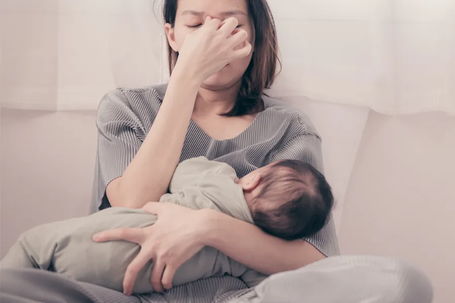 Postpartum-Hormone-Imbalance-Clinic A woman nursing an infant on couch rubs her eyes with exhaustion. Get treatment for postpartum hormone imbalance from Raving Health in Virginia Beach.
