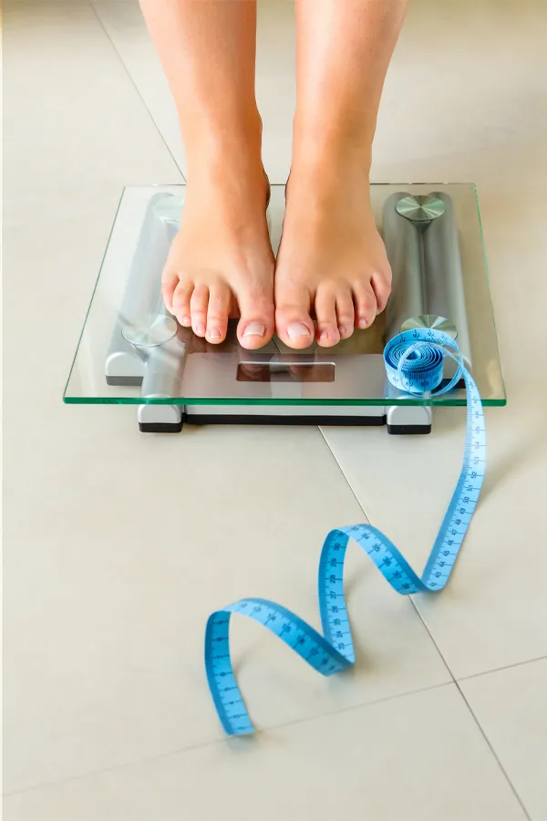 Weight-Loss-Resistance-Treatment Close-up of a woman's feet standing on a scale, with measuring tape by her toes, getting treatment for weight loss resistance from Raving Health in Virginia Beach.